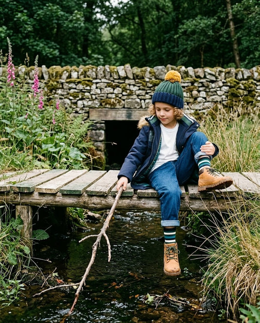 Niño con abrigo Outside azul marino sentado en un puente junto a un arroyo, estilo urbano y casual